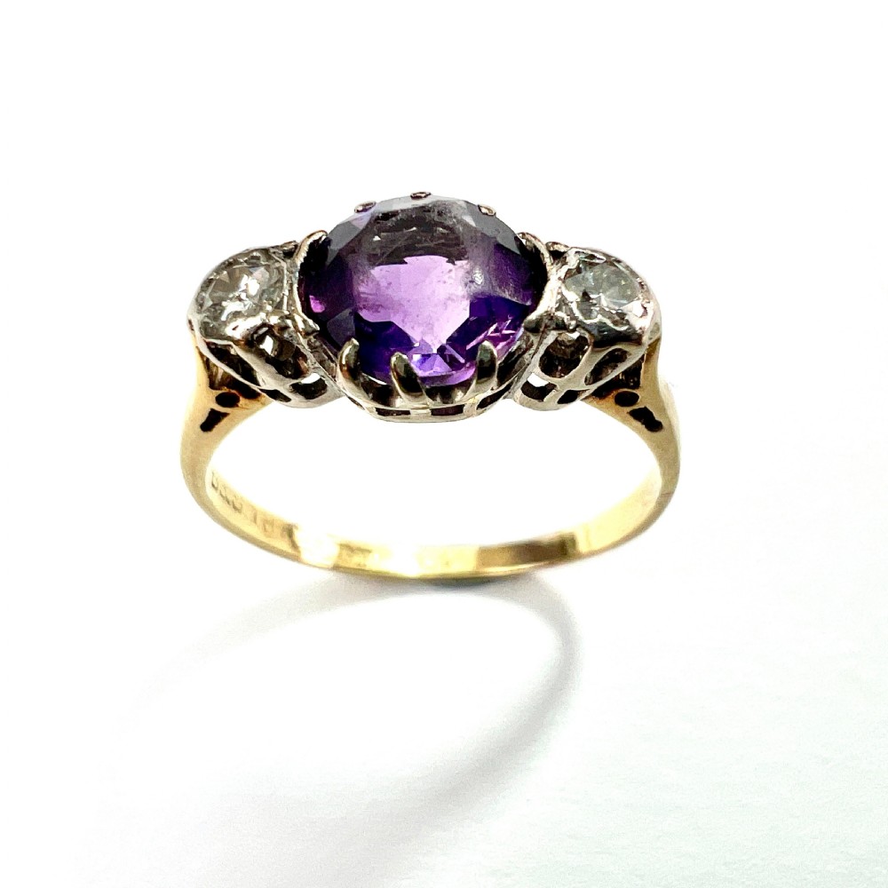 An antique 18ct yellow gold ring featuring a round purple central amethyst flanked by two diamonds in platinum settings. View X