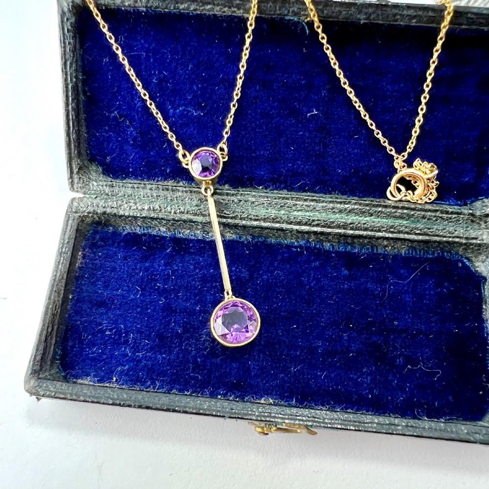 An Edwardian gold drop necklace featuring two round bezel-set purple amethyst stones connected by a slender gold bar on a fine chain.