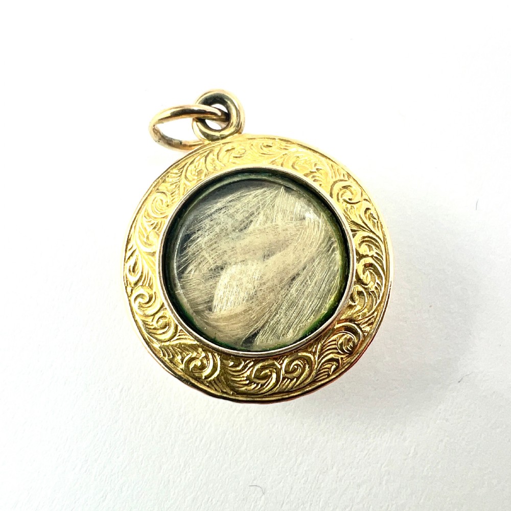 A circular 15ct gold Victorian locket pendant featuring hand-engraved scrollwork around a central domed glass window containing a lock of hair.