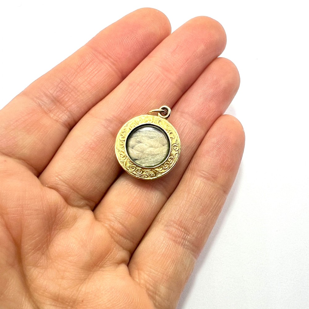 A circular 15ct gold Victorian locket pendant featuring hand-engraved scrollwork around a central domed glass window containing a lock of hair. View V