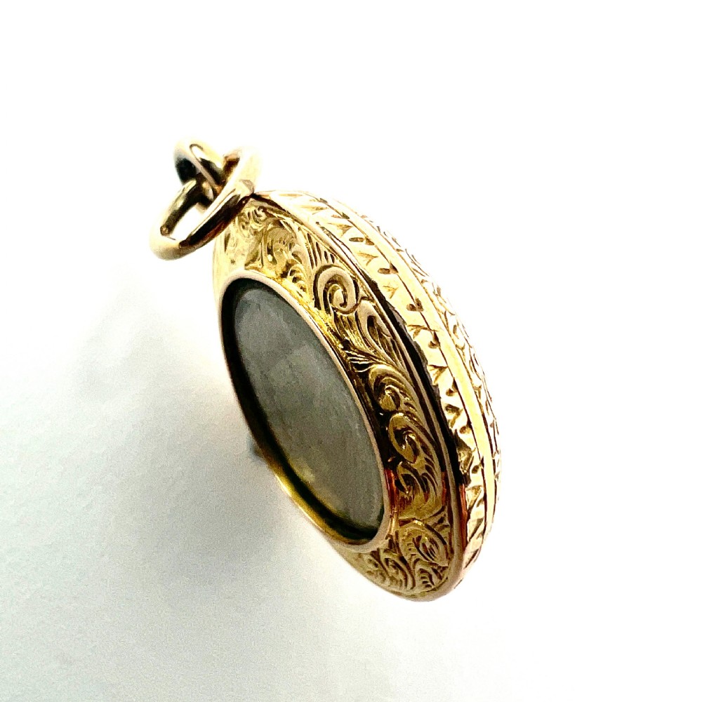 A circular 15ct gold Victorian locket pendant featuring hand-engraved scrollwork around a central domed glass window containing a lock of hair. View VI