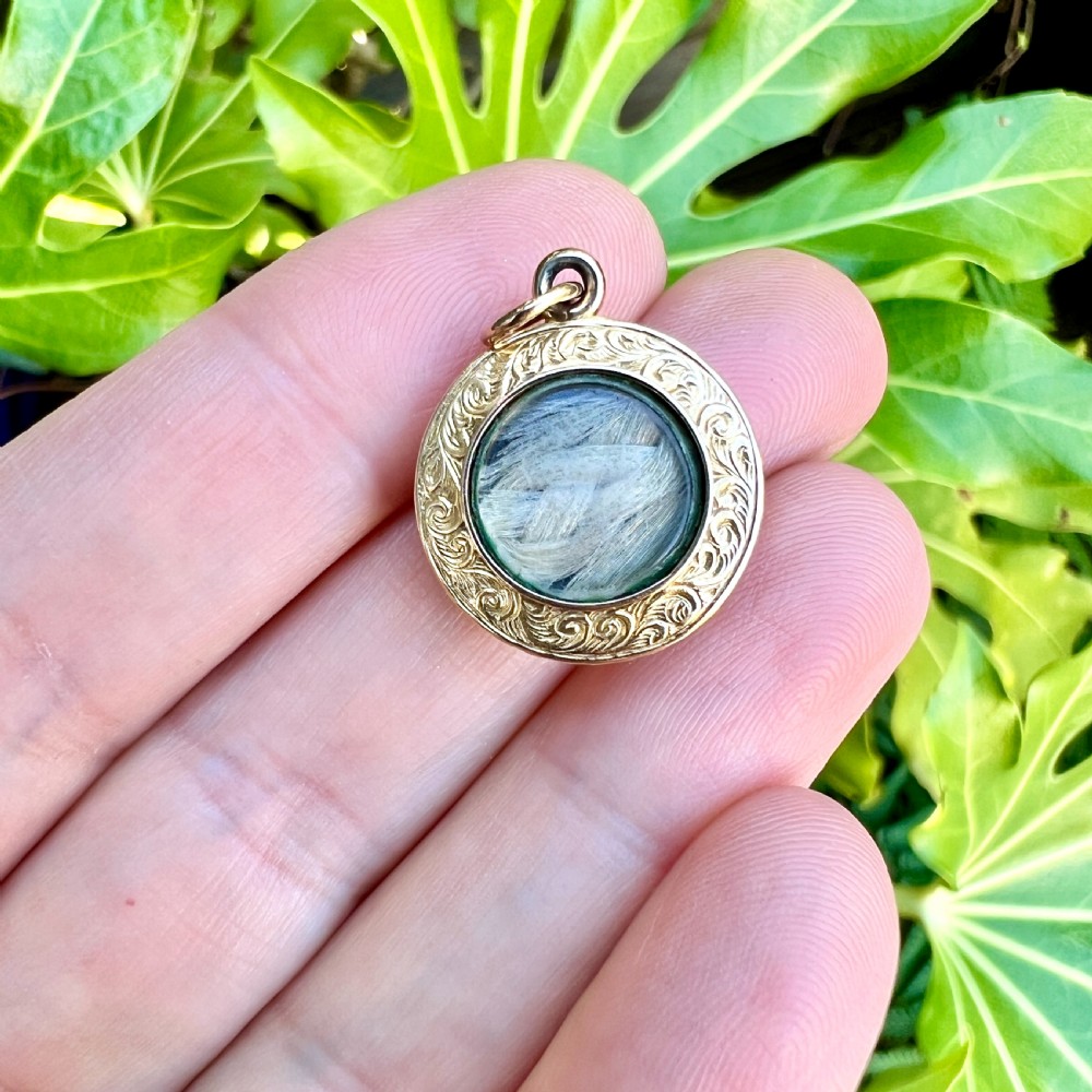 A circular 15ct gold Victorian locket pendant featuring hand-engraved scrollwork around a central domed glass window containing a lock of hair. View VIII