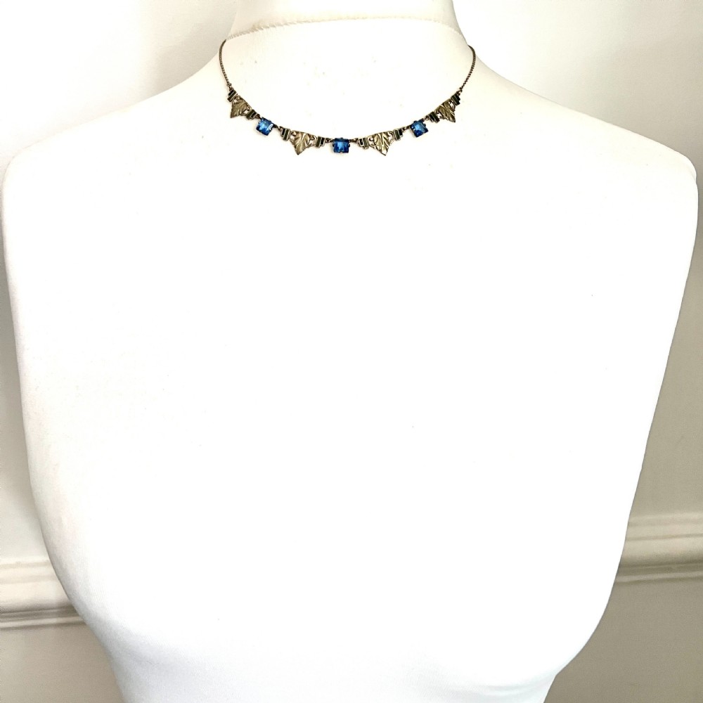 An art deco rolled gold necklace featuring square blue paste stones, leaf motifs, and dark enamel accents on a white background. View VI