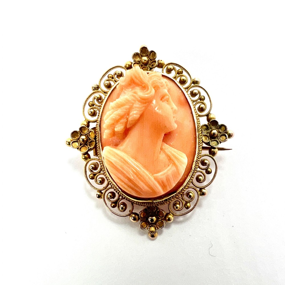 An oval Victorian brooch featuring a hand-carved peach coral classical female cameo profile set within a gold filigree wirework frame.
