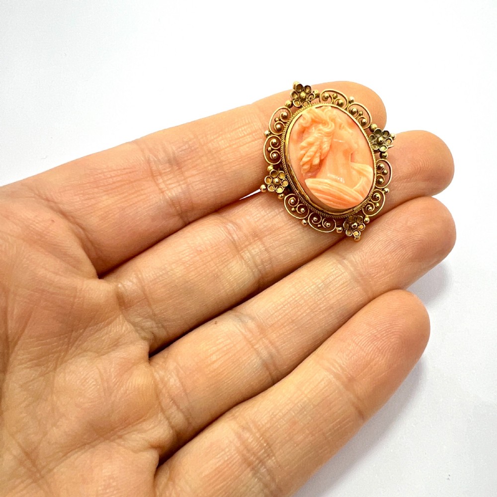 An oval Victorian brooch featuring a hand-carved peach coral classical female cameo profile set within a gold filigree wirework frame. View V