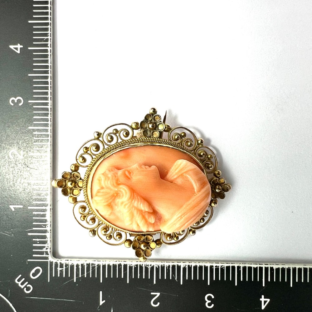 An oval Victorian brooch featuring a hand-carved peach coral classical female cameo profile set within a gold filigree wirework frame. View VI
