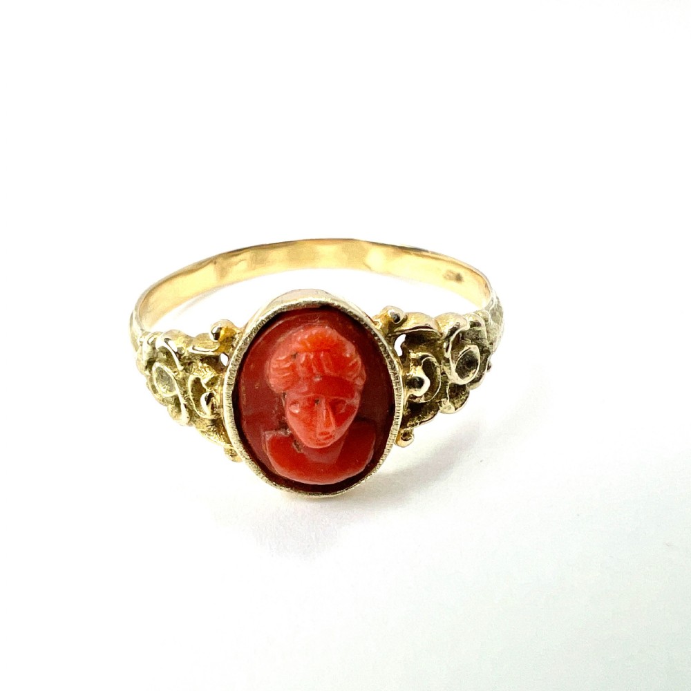 An antique gold ring featuring a central oval carved red coral cameo depicting a classical profile with decorative scrollwork shoulders.