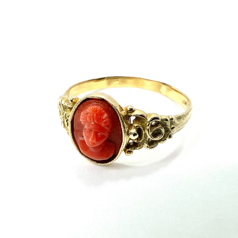 An antique gold ring featuring a central oval carved red coral cameo depicting a classical profile with decorative scrollwork shoulders. View II