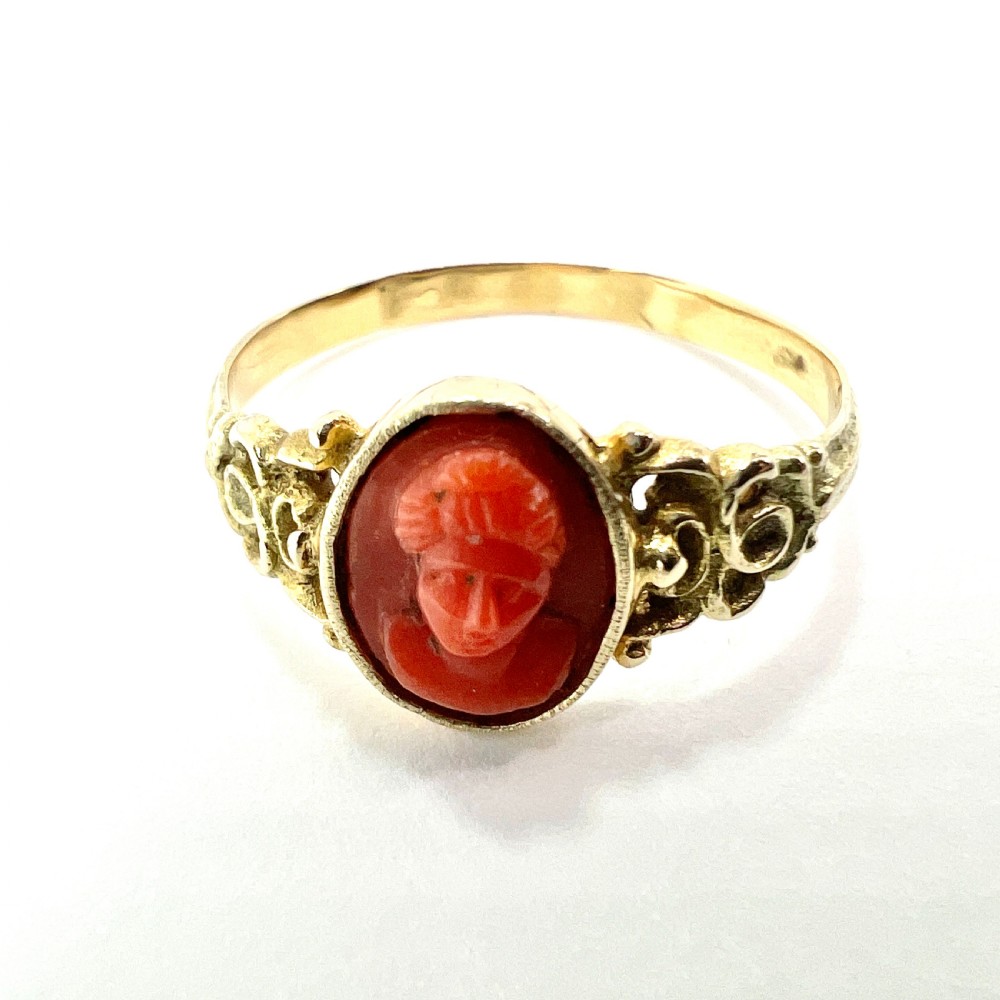 An antique gold ring featuring a central oval carved red coral cameo depicting a classical profile with decorative scrollwork shoulders. View III