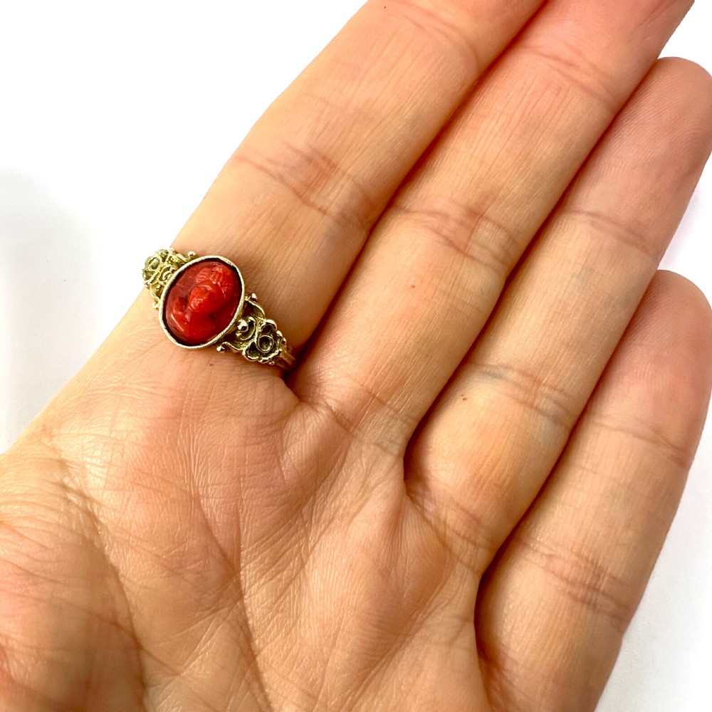 An antique gold ring featuring a central oval carved red coral cameo depicting a classical profile with decorative scrollwork shoulders. View VIII