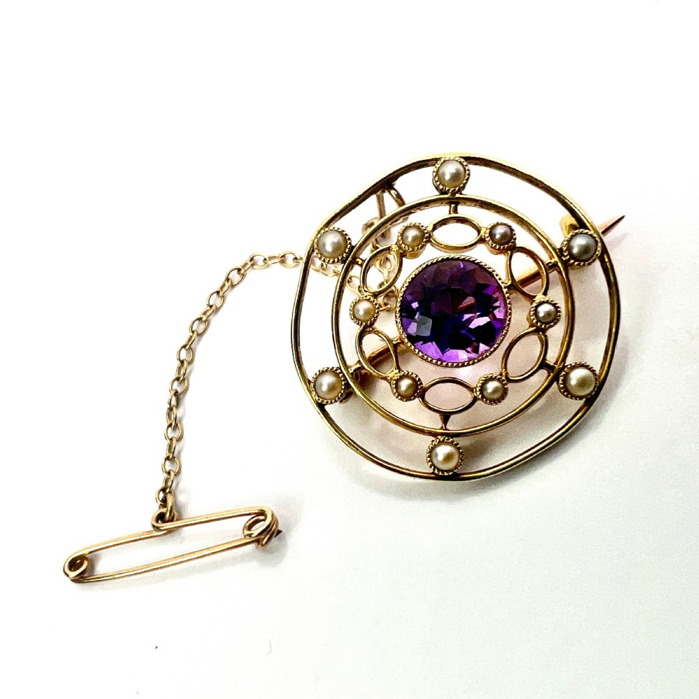 An Edwardian 9ct yellow gold circular target brooch featuring a central round purple amethyst surrounded by openwork concentric rings set with seed pearls, attached to a safety chain.