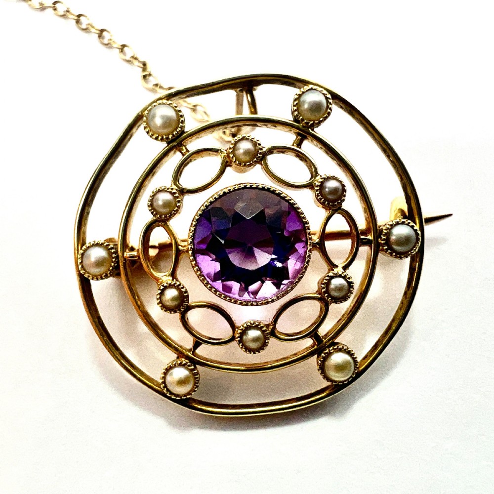 An Edwardian 9ct yellow gold circular target brooch featuring a central round purple amethyst surrounded by openwork concentric rings set with seed pearls, attached to a safety chain. View II
