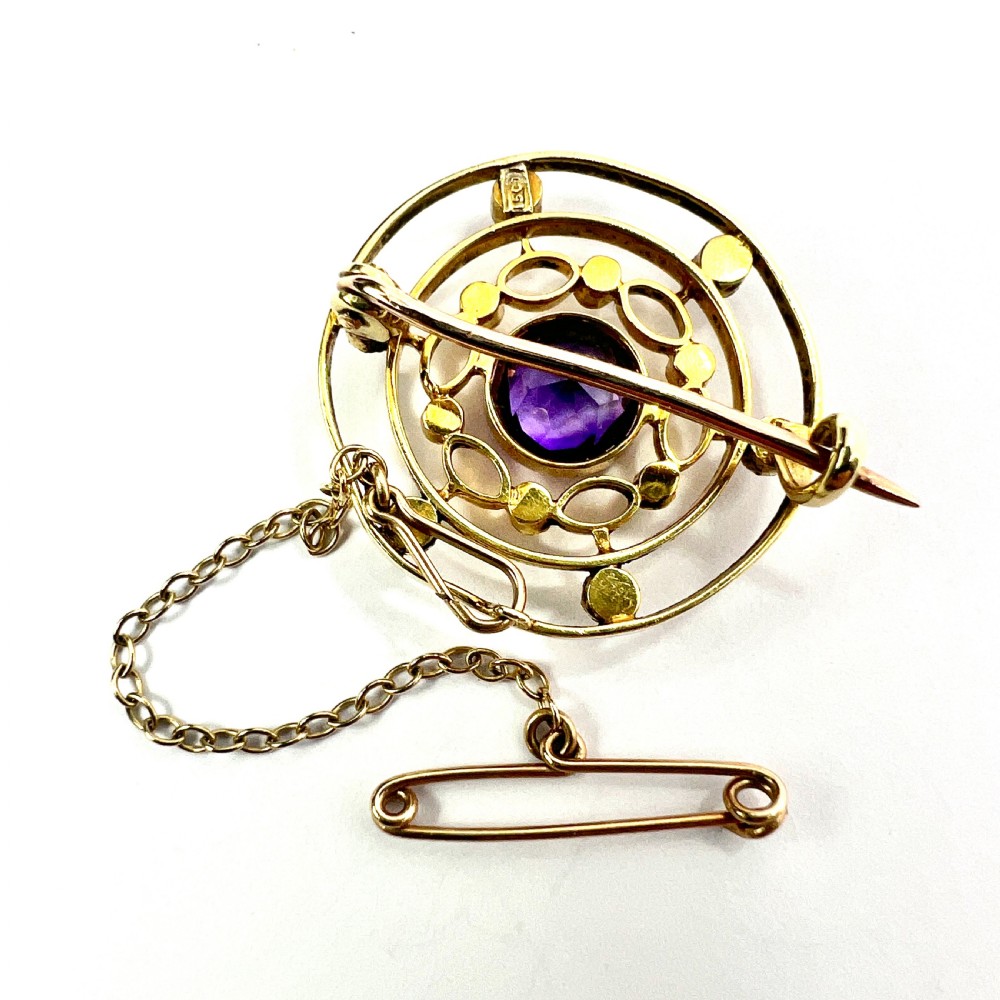 An Edwardian 9ct yellow gold circular target brooch featuring a central round purple amethyst surrounded by openwork concentric rings set with seed pearls, attached to a safety chain. View III