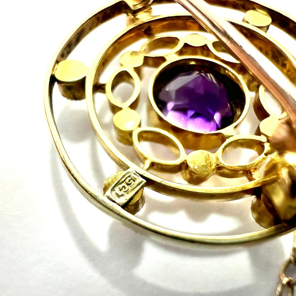 An Edwardian 9ct yellow gold circular target brooch featuring a central round purple amethyst surrounded by openwork concentric rings set with seed pearls, attached to a safety chain. View IV