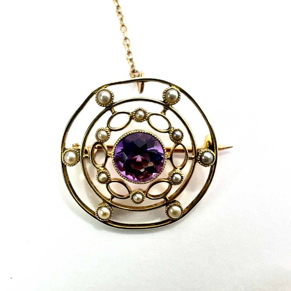 An Edwardian 9ct yellow gold circular target brooch featuring a central round purple amethyst surrounded by openwork concentric rings set with seed pearls, attached to a safety chain. View V