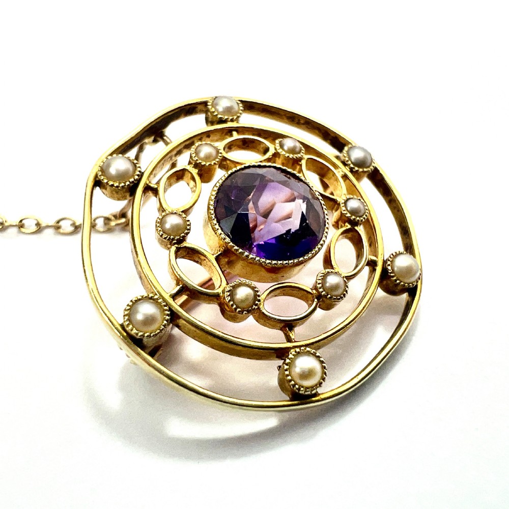 An Edwardian 9ct yellow gold circular target brooch featuring a central round purple amethyst surrounded by openwork concentric rings set with seed pearls, attached to a safety chain. View VI