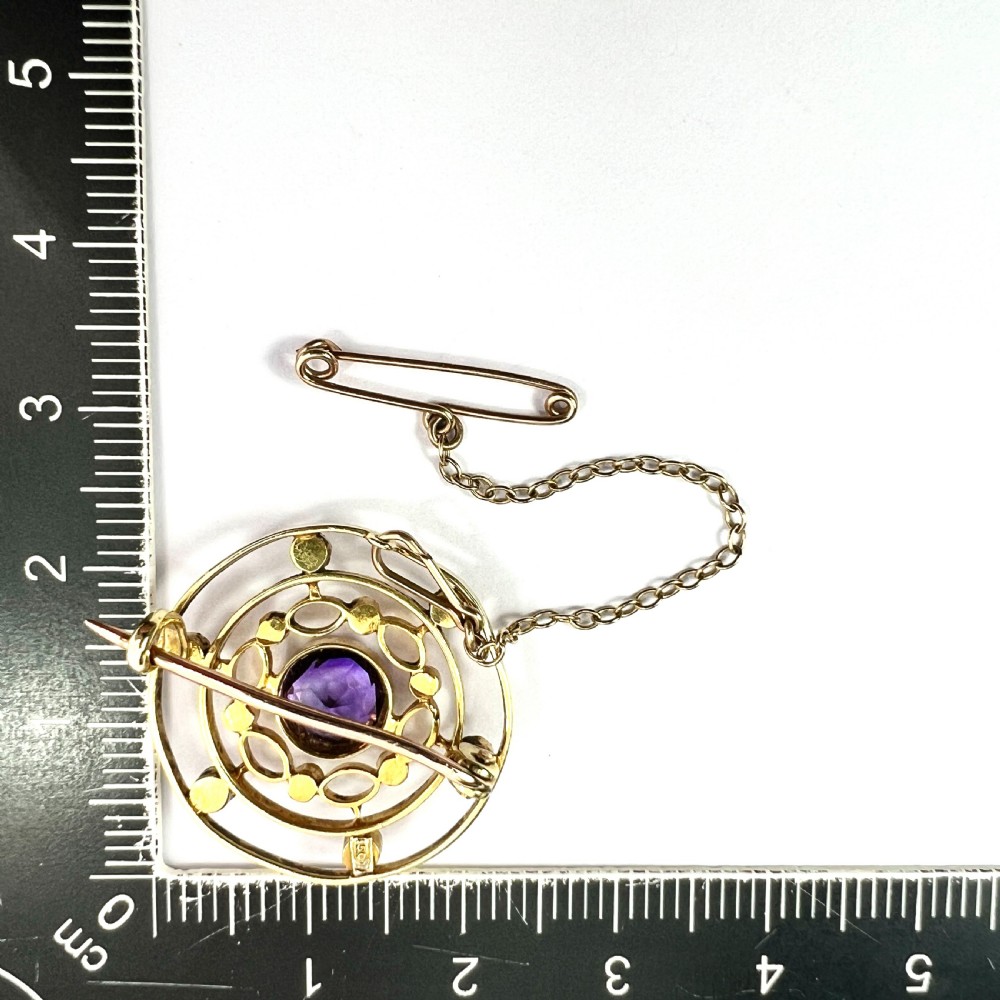 An Edwardian 9ct yellow gold circular target brooch featuring a central round purple amethyst surrounded by openwork concentric rings set with seed pearls, attached to a safety chain. View VII