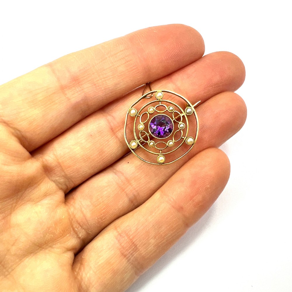 An Edwardian 9ct yellow gold circular target brooch featuring a central round purple amethyst surrounded by openwork concentric rings set with seed pearls, attached to a safety chain. View VIII