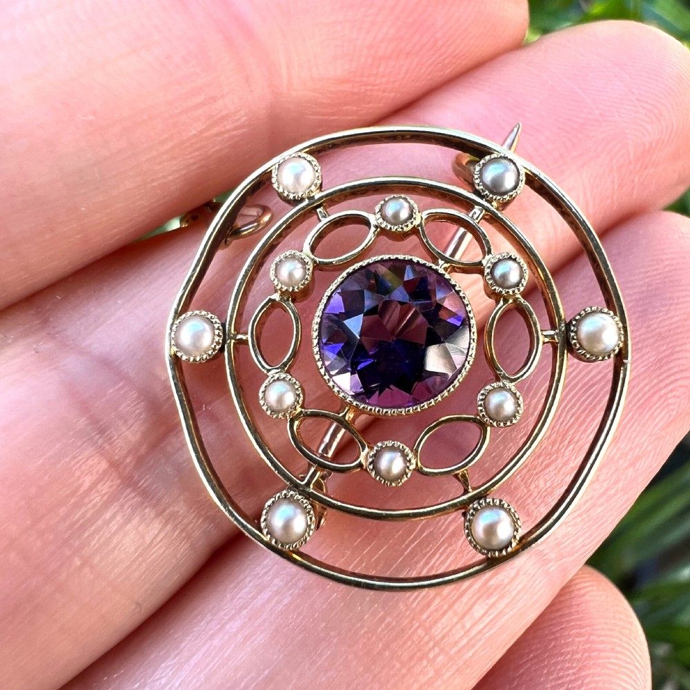 An Edwardian 9ct yellow gold circular target brooch featuring a central round purple amethyst surrounded by openwork concentric rings set with seed pearls, attached to a safety chain. View IX