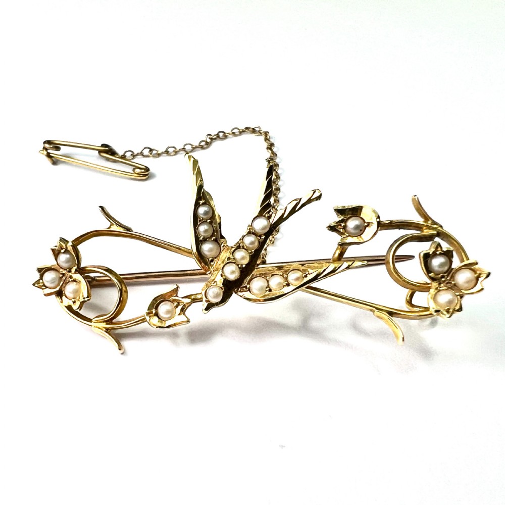 An Edwardian 15ct yellow gold brooch featuring a central swallow motif set with seed pearls alongside floral scrollwork and a safety chain attachment.
