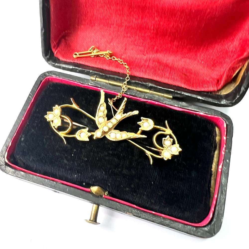 An Edwardian 15ct yellow gold brooch featuring a central swallow motif set with seed pearls alongside floral scrollwork and a safety chain attachment. View II