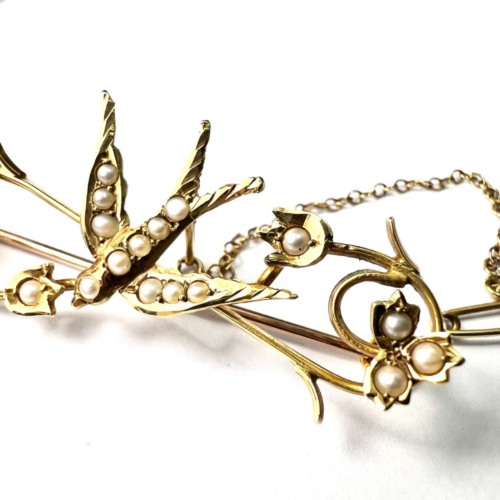 An Edwardian 15ct yellow gold brooch featuring a central swallow motif set with seed pearls alongside floral scrollwork and a safety chain attachment. View V