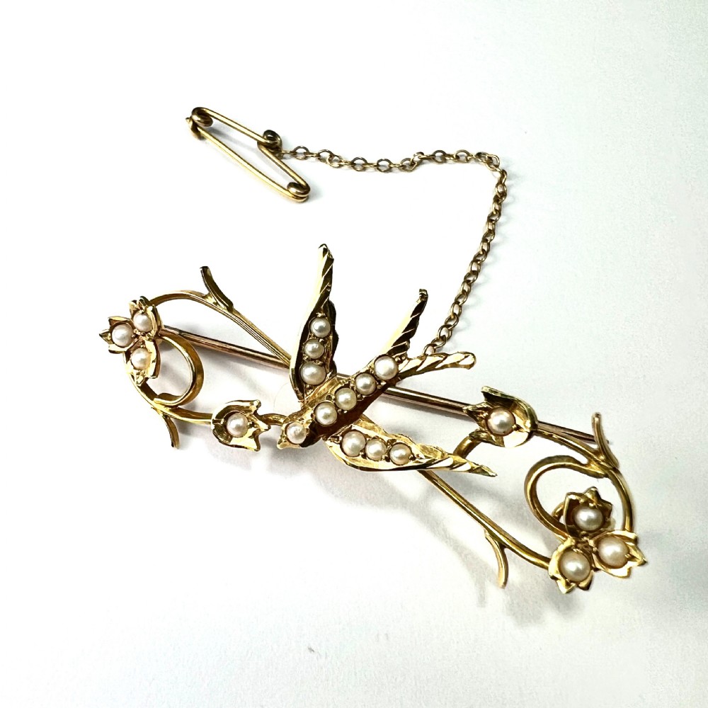 An Edwardian 15ct yellow gold brooch featuring a central swallow motif set with seed pearls alongside floral scrollwork and a safety chain attachment. View VI
