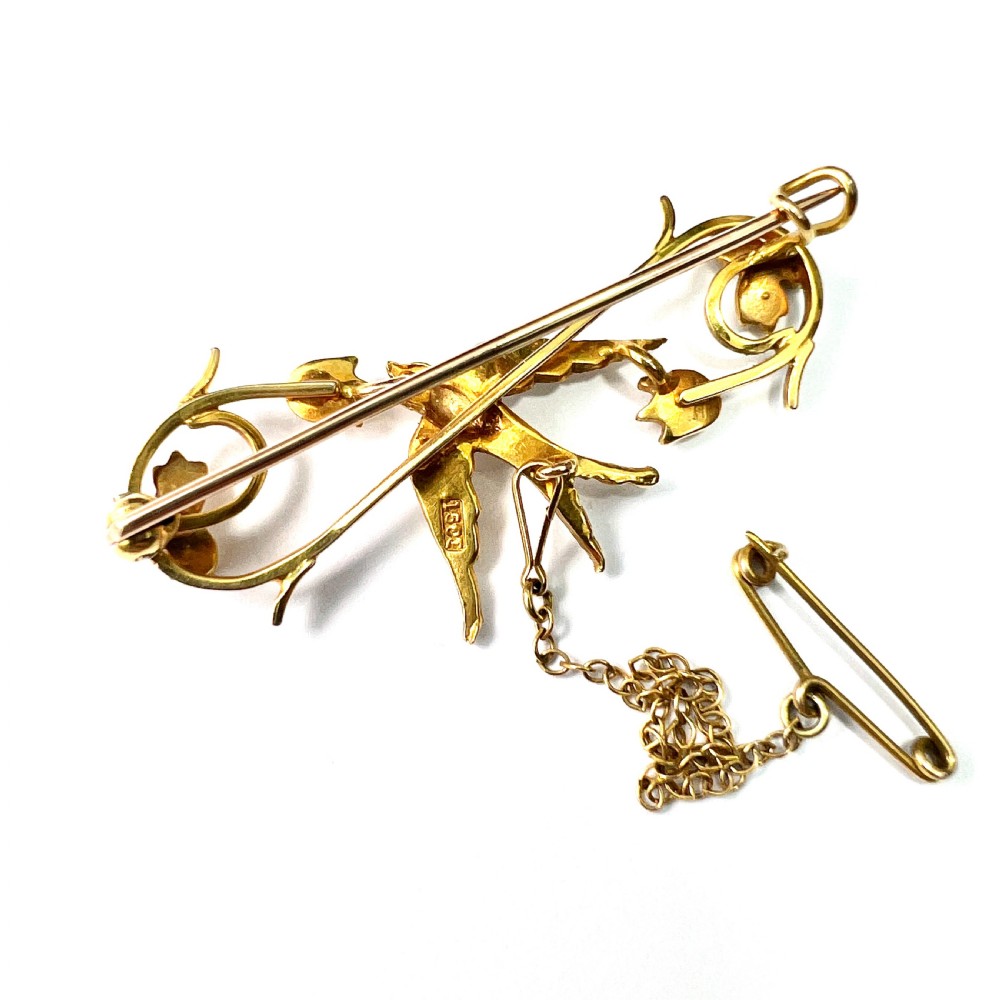 An Edwardian 15ct yellow gold brooch featuring a central swallow motif set with seed pearls alongside floral scrollwork and a safety chain attachment. View VII