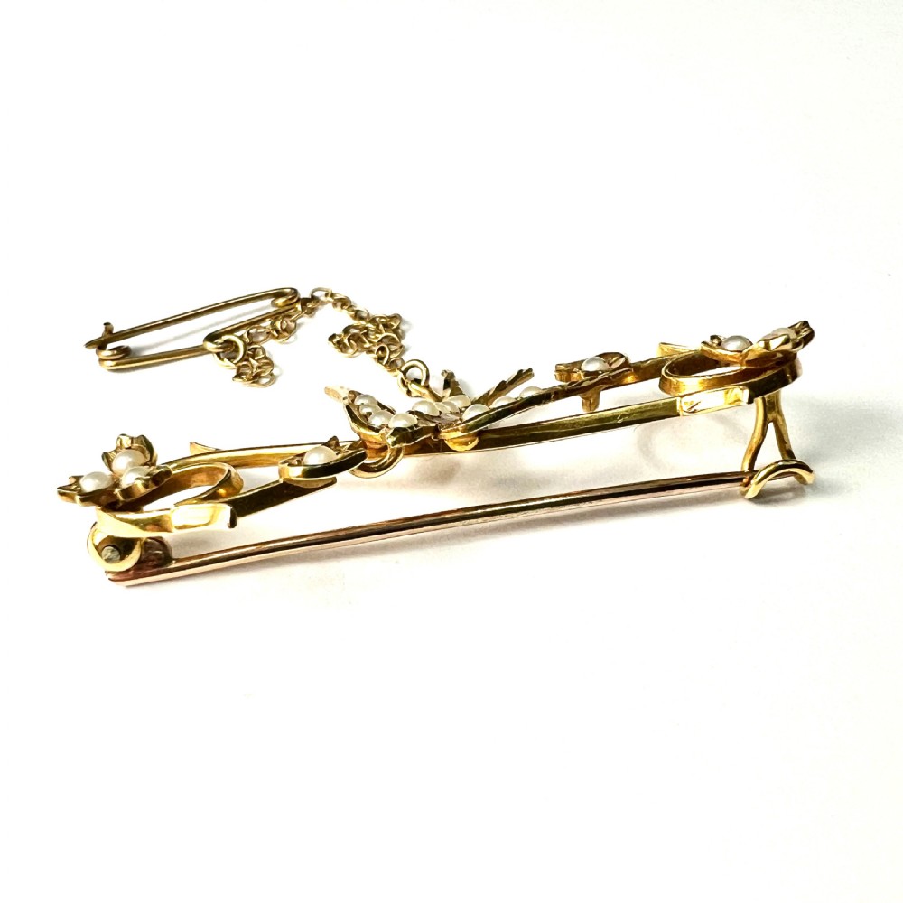 An Edwardian 15ct yellow gold brooch featuring a central swallow motif set with seed pearls alongside floral scrollwork and a safety chain attachment. View VIII