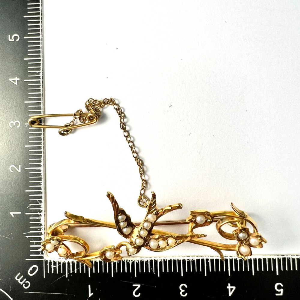An Edwardian 15ct yellow gold brooch featuring a central swallow motif set with seed pearls alongside floral scrollwork and a safety chain attachment. View IX