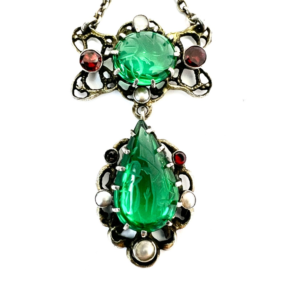 An antique silver pendant featuring carved green paste stones, round garnets, and seed pearls set in an openwork filigree frame.