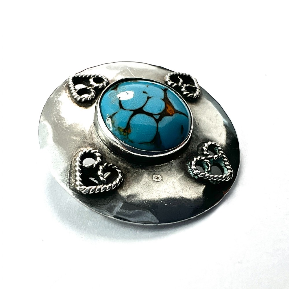 A circular hand-hammered silver brooch featuring a central turquoise glass cabochon framed by four rope-twist heart motifs. View III