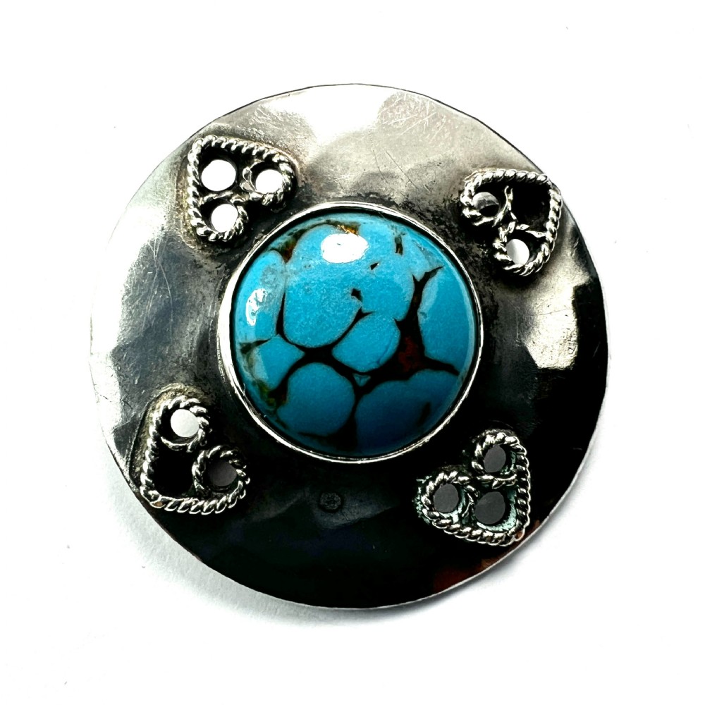 A circular hand-hammered silver brooch featuring a central turquoise glass cabochon framed by four rope-twist heart motifs. View V