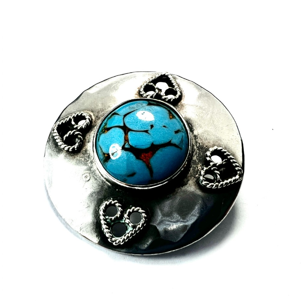 A circular hand-hammered silver brooch featuring a central turquoise glass cabochon framed by four rope-twist heart motifs. View VI