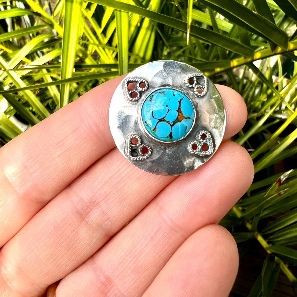 A circular hand-hammered silver brooch featuring a central turquoise glass cabochon framed by four rope-twist heart motifs. View VII