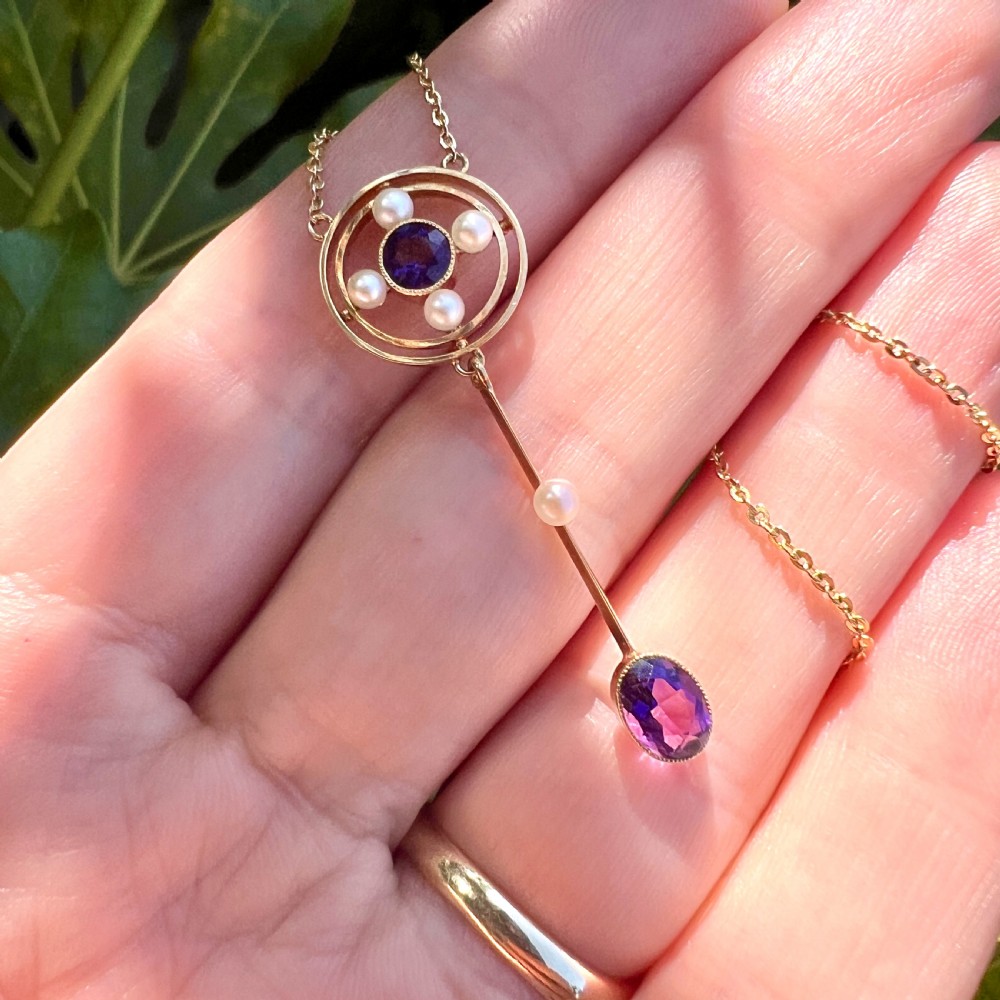 An Edwardian 15ct gold chain necklace featuring a circular pendant set with a round amethyst and four pearls, with a linear drop holding a seed pearl and an oval facet-cut amethyst. View VIII