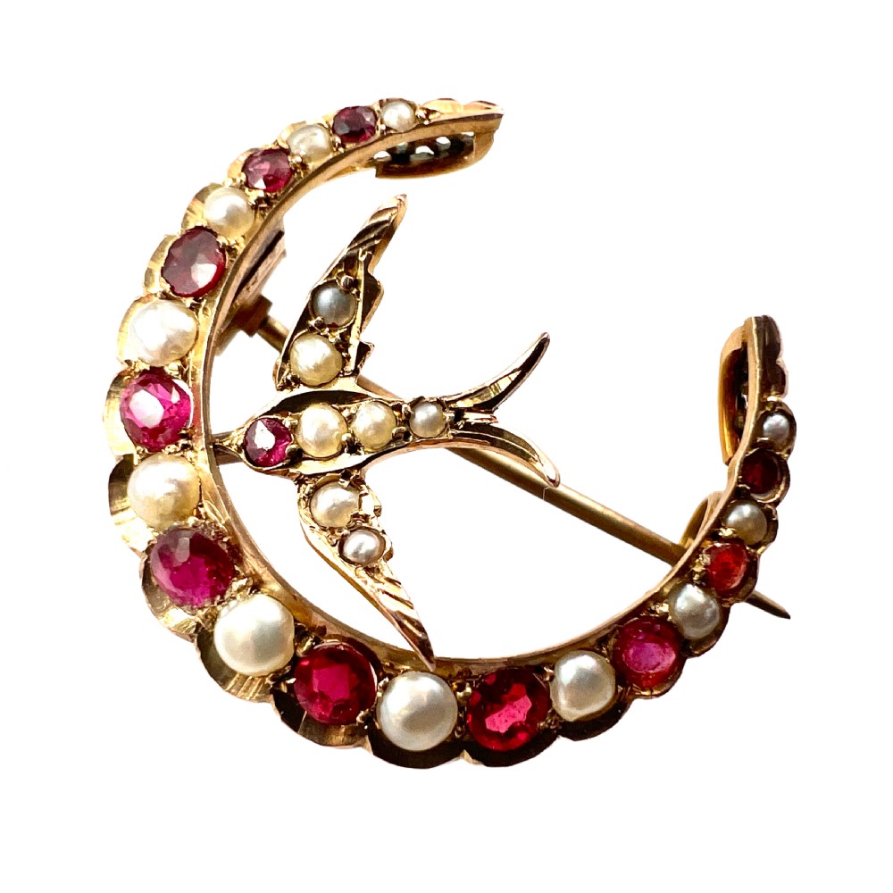A 9ct gold crescent moon brooch set with alternating rubies and seed pearls surrounding a central swallow motif embellished with pearls and a ruby eye.