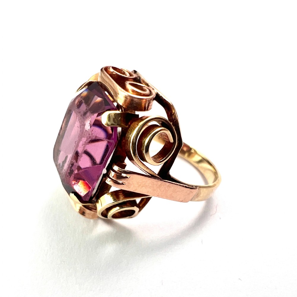 A 1940s Retro 14ct gold cocktail ring featuring a large rectangular cut purple paste gemstone set within ornate metal scrollwork.