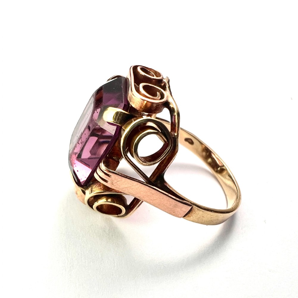 A 1940s Retro 14ct gold cocktail ring featuring a large rectangular cut purple paste gemstone set within ornate metal scrollwork. View IV