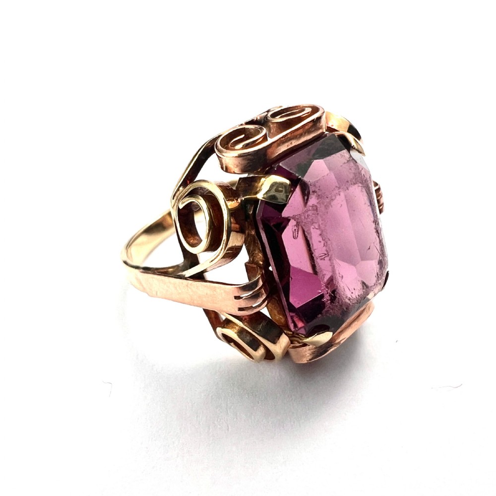 A 1940s Retro 14ct gold cocktail ring featuring a large rectangular cut purple paste gemstone set within ornate metal scrollwork. View VII