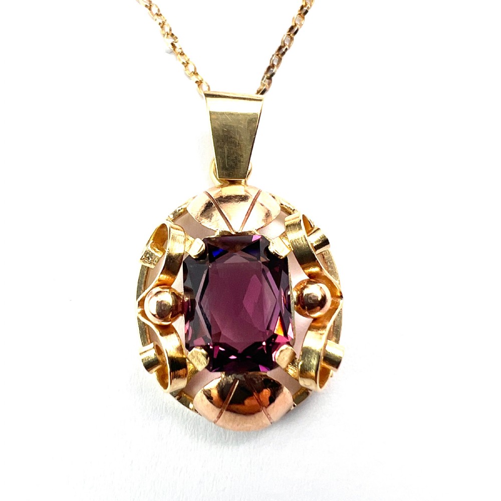 A 14ct yellow gold 1940s pendant featuring a central emerald-cut purple paste stone framed by sculptural gold scrolls and sphere accents. View V