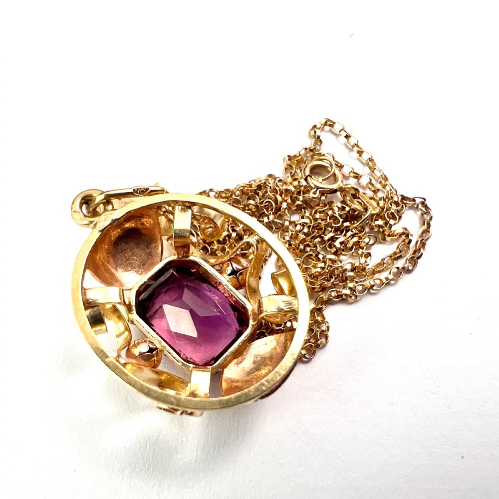 A 14ct yellow gold 1940s pendant featuring a central emerald-cut purple paste stone framed by sculptural gold scrolls and sphere accents. View VI
