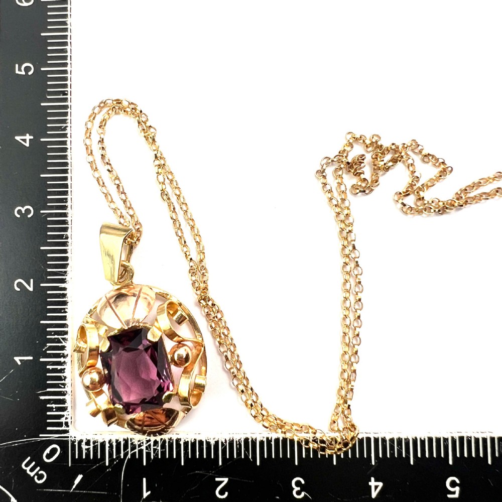 A 14ct yellow gold 1940s pendant featuring a central emerald-cut purple paste stone framed by sculptural gold scrolls and sphere accents. View IX
