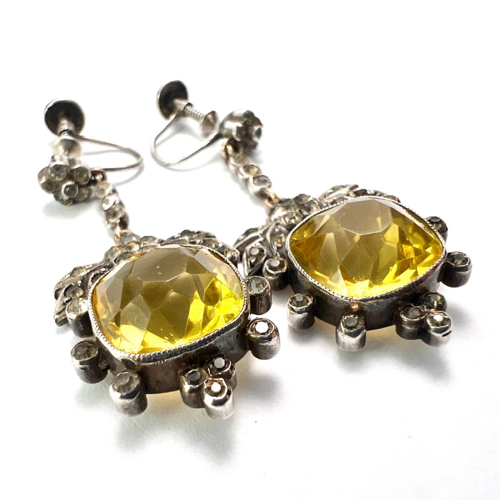 A pair of Victorian silver drop earrings featuring cushion-cut yellow citrine paste stones surrounded by foliate settings with clear glass accents. View V