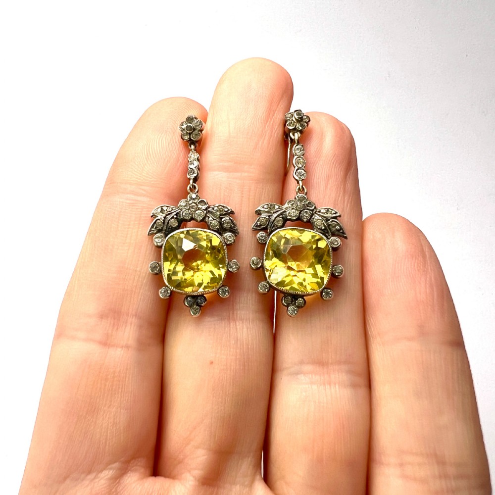 A pair of Victorian silver drop earrings featuring cushion-cut yellow citrine paste stones surrounded by foliate settings with clear glass accents. View X