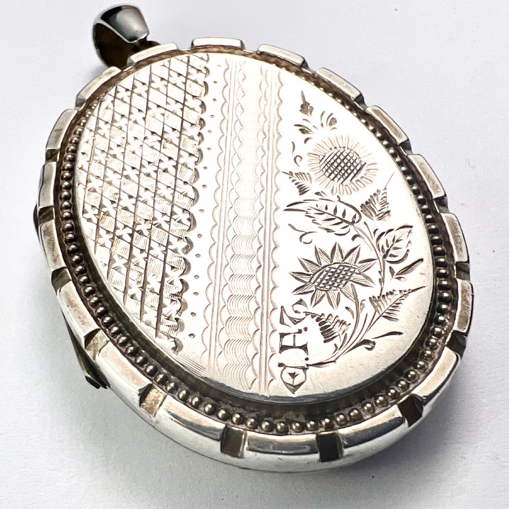 An oval Victorian silver locket decorated with intricate floral engraving, geometric crosshatching, and a beaded border.