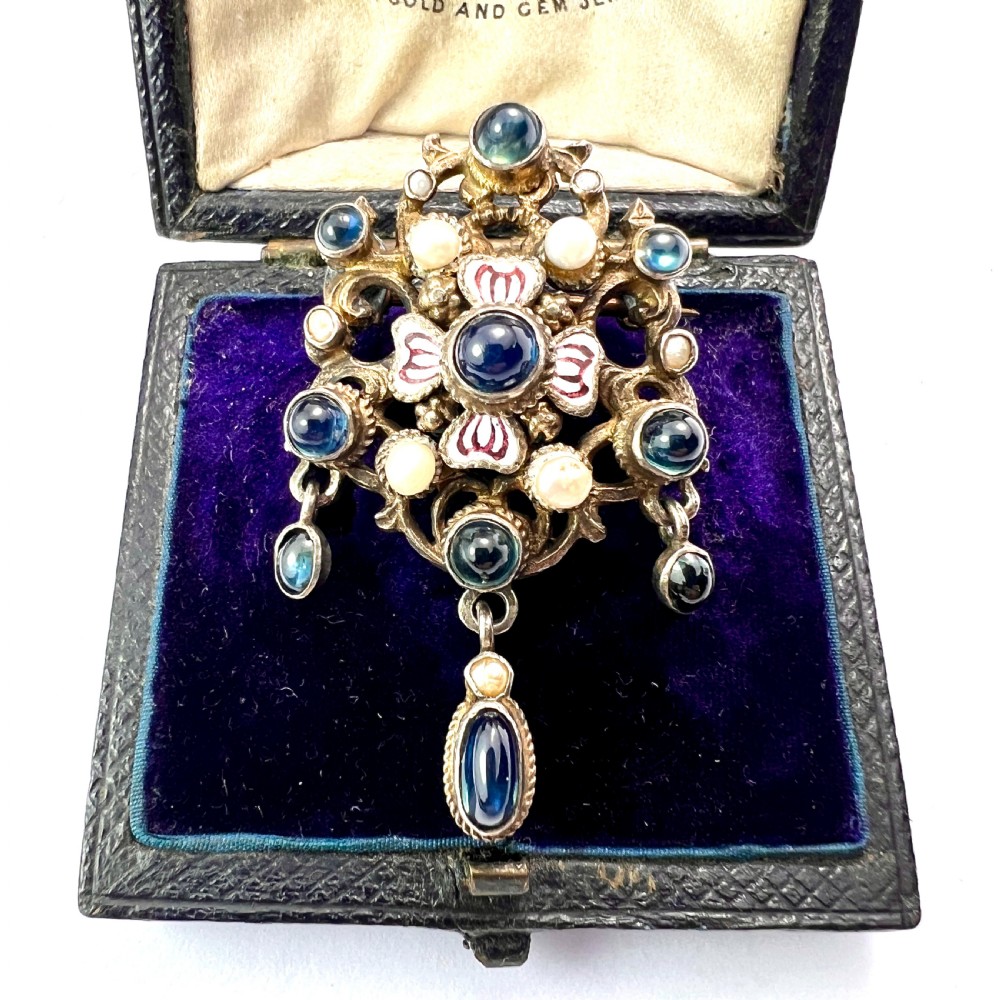 An Austro-Hungarian silver-gilt brooch decorated with blue cabochons, seed pearls, polychrome enamel, and a dangling teardrop pendant.