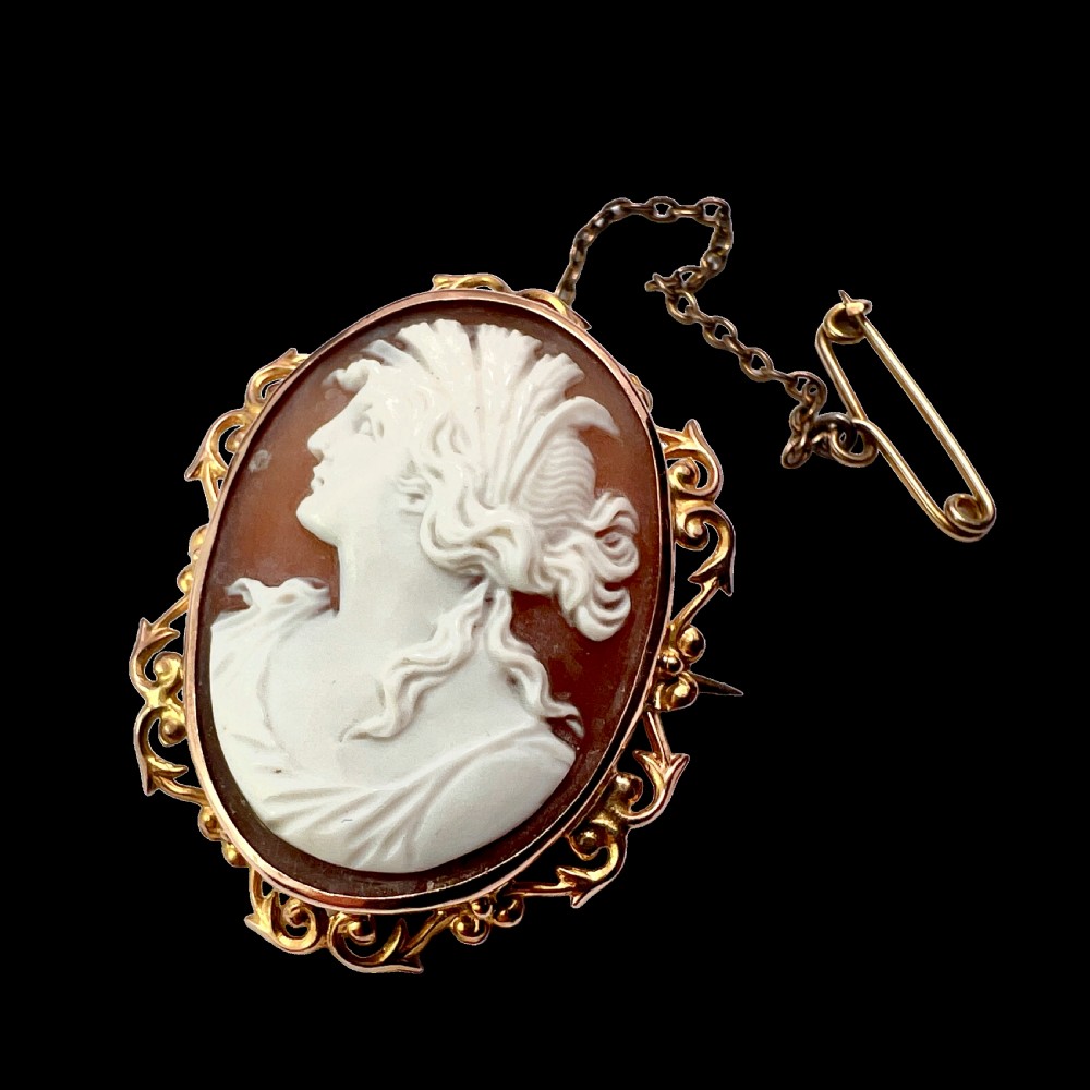 An oval Victorian 9ct gold brooch featuring a white carved shell cameo of a classical female profile set within an ornate openwork scroll frame with an attached safety pin.
