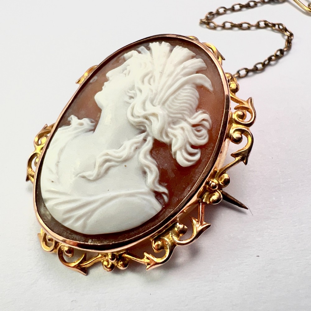 An oval Victorian 9ct gold brooch featuring a white carved shell cameo of a classical female profile set within an ornate openwork scroll frame with an attached safety pin. View V
