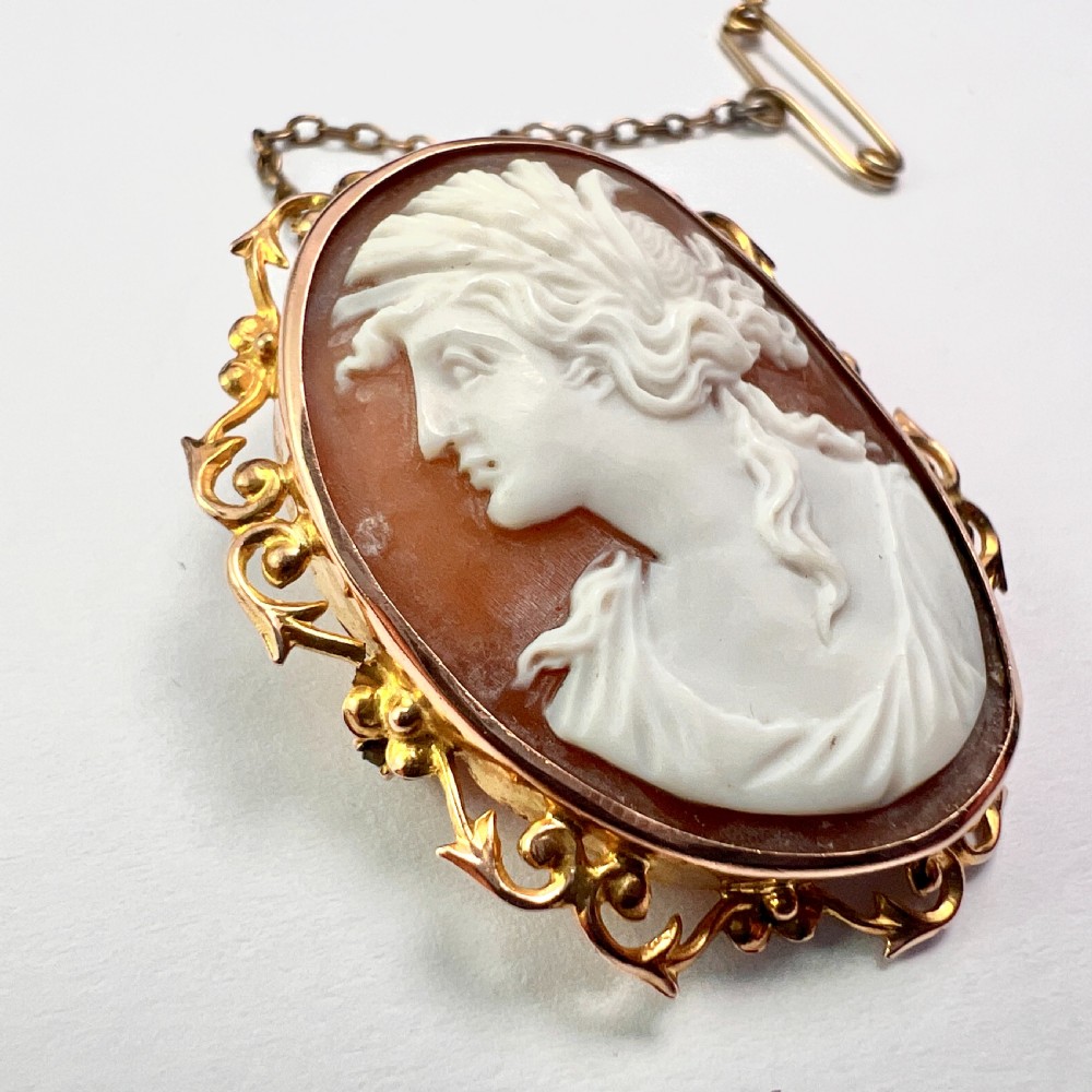An oval Victorian 9ct gold brooch featuring a white carved shell cameo of a classical female profile set within an ornate openwork scroll frame with an attached safety pin. View VI
